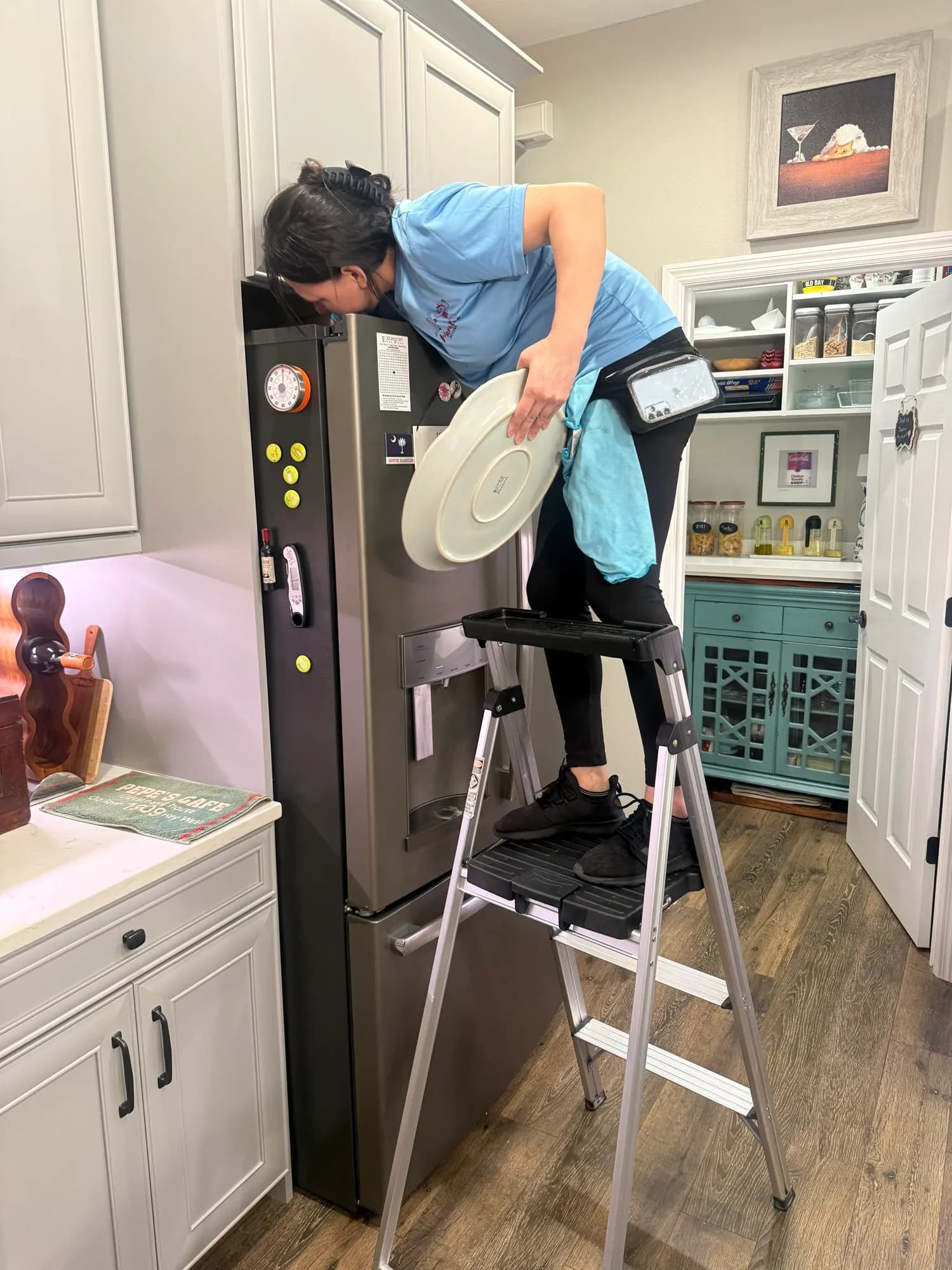 Cleaner on a step ladder wiping the top of a refrigerator in an upscale kitchen