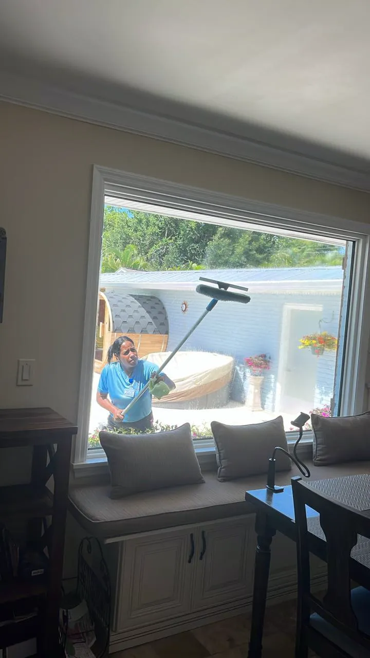 Cleaner washing a large picture window, viewed from a styled living room with a window seat